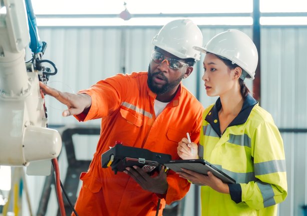 Man and woman wearing hard hats and coveralls inspecting a robotic arm for safety