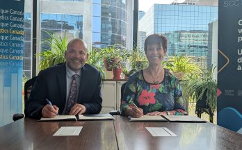 André Loranger (left) and Chantal Guay (right) sitting at a table in front of a window, smiling and signing documents. Behind them are a branded banner for Statistics Canada and the Standards Council of Canada 