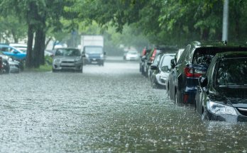 Rue urbaine inondée avec des voitures garées partiellement submergées dans l’eau, forte pluie et arbres verdoyants bordant la route