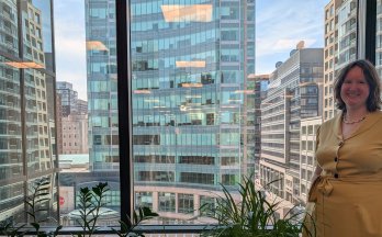 Smiling woman standing by an office window with tall buildings in the background.
