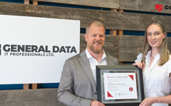 Two people holding a framed certificate in front of a General Data IT Professionals Ltd. sign.