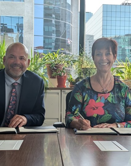 André Loranger (left) and Chantal Guay (right) sitting at a table in front of a window, smiling and signing documents. Behind them are a branded banner for Statistics Canada and the Standards Council of Canada 