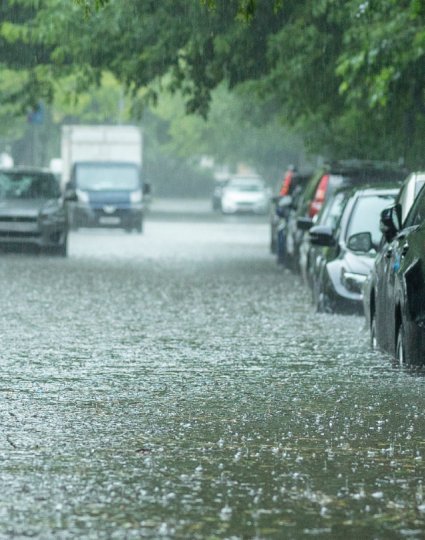 Rue urbaine inondée avec des voitures garées partiellement submergées dans l’eau, forte pluie et arbres verdoyants bordant la route