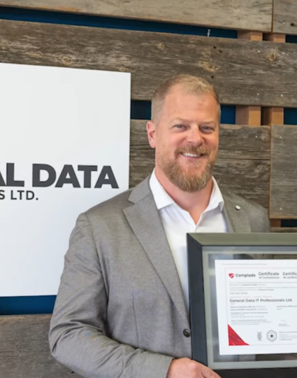 Two people holding a framed certificate in front of a General Data IT Professionals Ltd. sign.