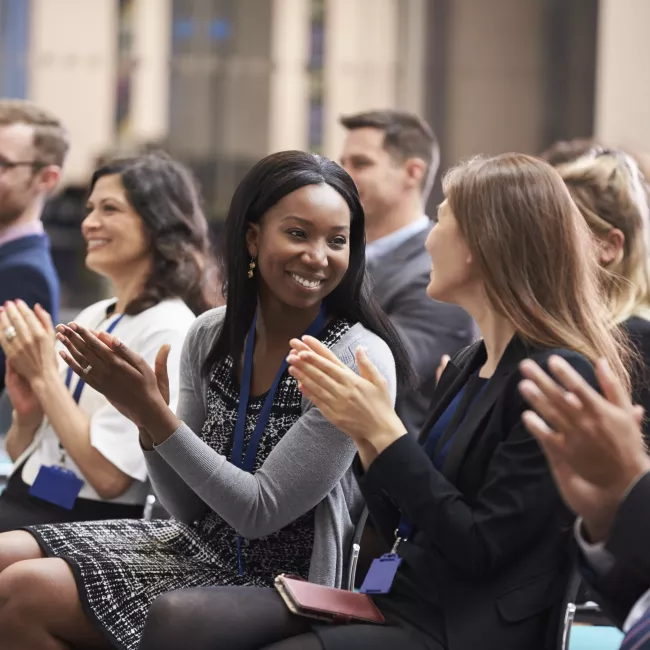 Diverse group of professionals in an audience applauding speaker after conference presentation.