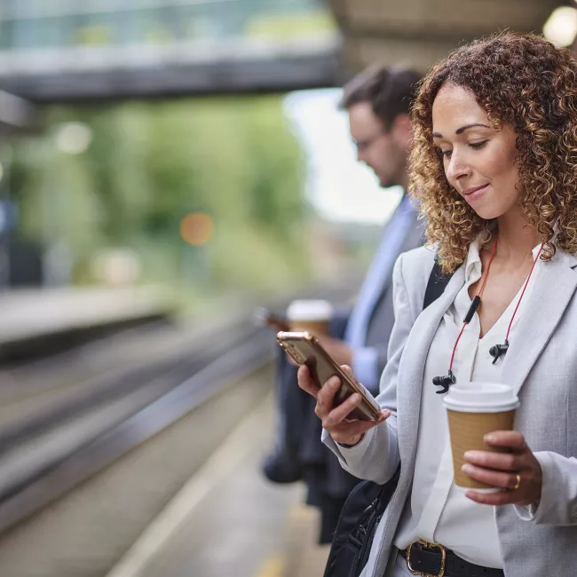 Businesswomen waiting for train on her morning commute looking at her phone holding a cup of coffee.