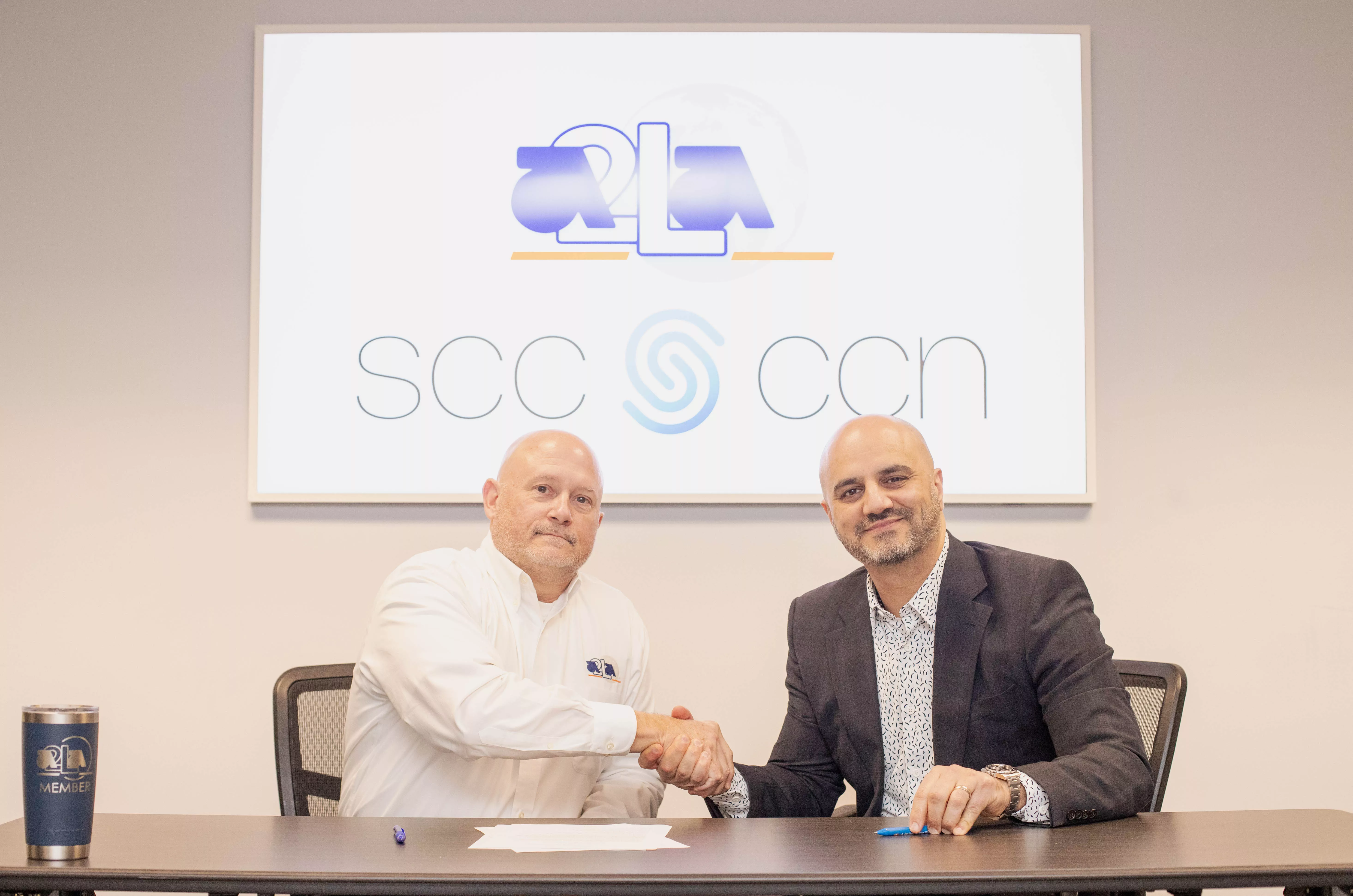  Two men shaking hands at a table, with SCC and A2LA logos displayed on the screen behind them