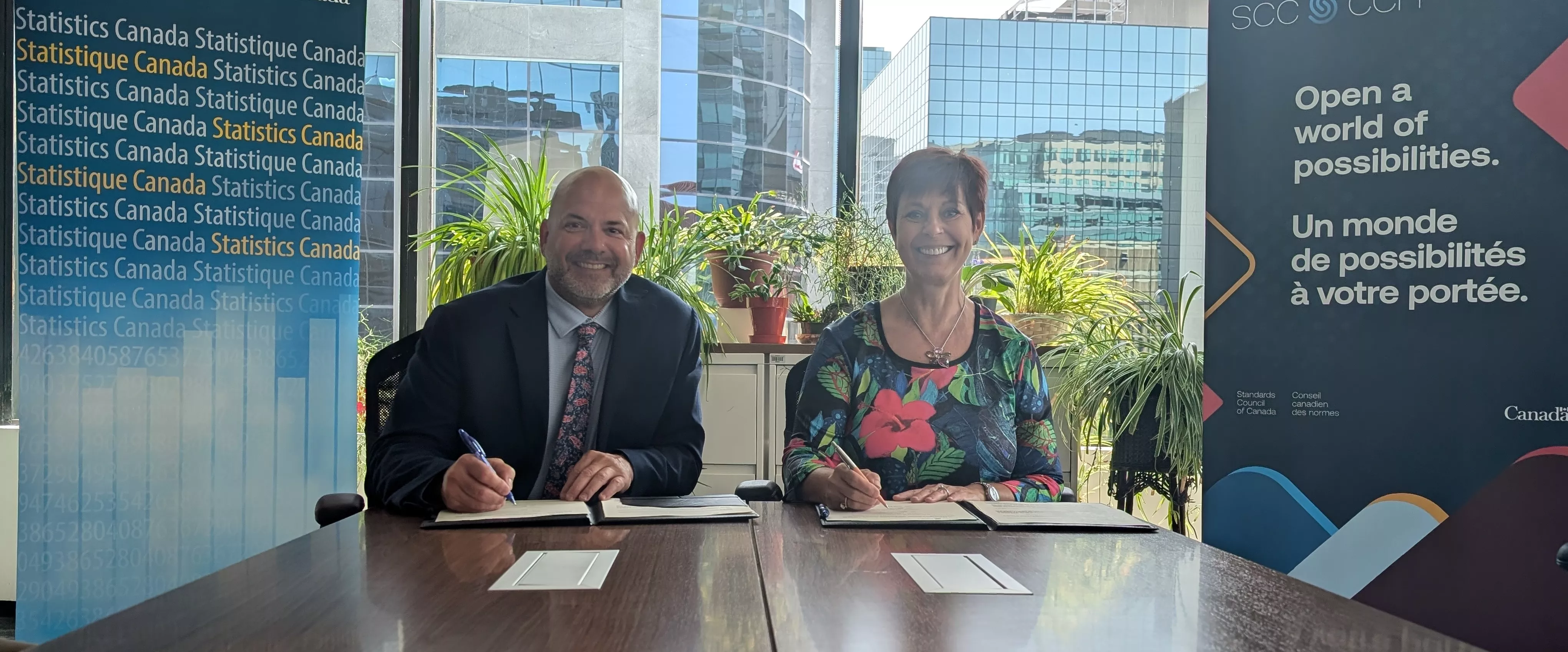 André Loranger (left) and Chantal Guay (right) sitting at a table in front of a window, smiling and signing documents. Behind them are a branded banner for Statistics Canada and the Standards Council of Canada 