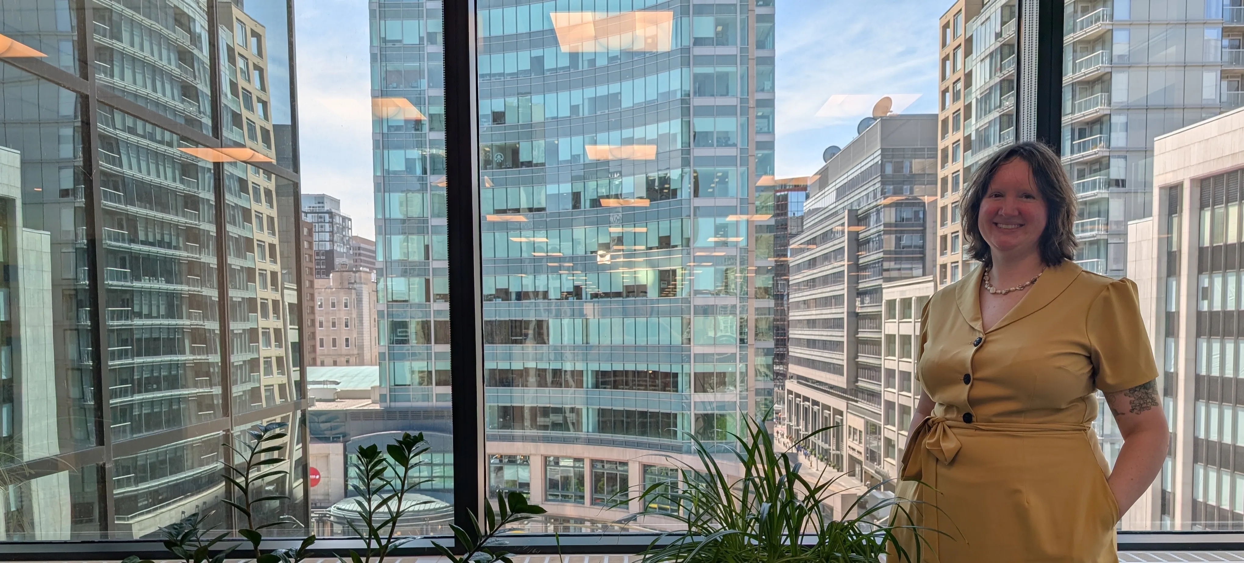 Smiling woman standing by an office window with tall buildings in the background.